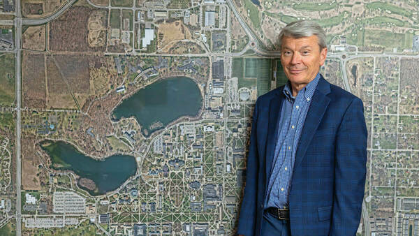 A man with gray hair wearing a light blue shirt and darker blue suit stands in front of a large map of the campus of Notre Dame.