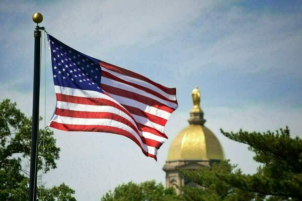 A United States flag waves in the foreground, with the Golden Dome of the Main Building at the University of Notre Dame blurred in the background.