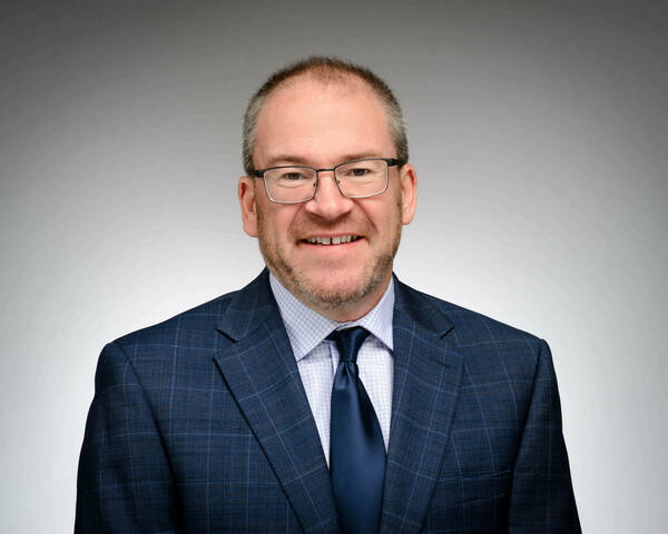 White man with glasses wearing a blue suit and tie, smiling in front of a gray background.