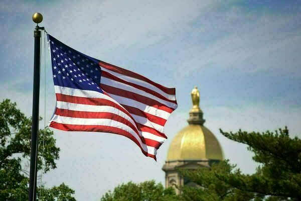 A United States flag waves in the foreground, with the Golden Dome of the Main Building at the University of Notre Dame blurred in the background.