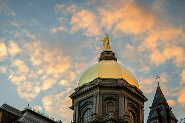 The Golden Dome of the Main Building at the University of Notre Dame, topped with the statue of Mary, against a sunset sky with orange and pink clouds.