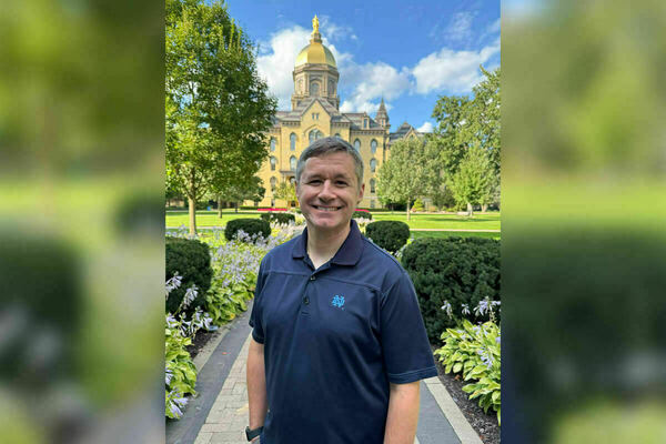 A man with grey hair and blue polo shirt stands in front of the Main Building, Golden Dome on the campus of Notre Dame