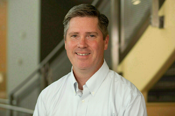 Smiling man with short graying hair in a light blue and white plaid shirt, in a modern building interior.