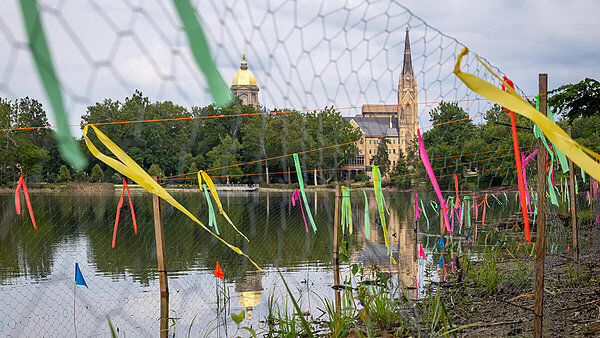 Colorful ribbons tied to a fence by a lake reflect Notre Dame's Golden Dome and Basilica of the Sacred Heart.